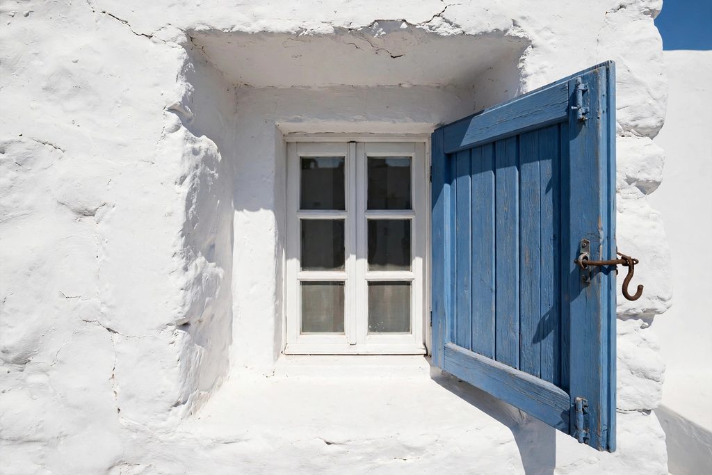 A small white-framed window set deep inside a thick, whitewashed stone wall with open blue shutters under bright sunlight.