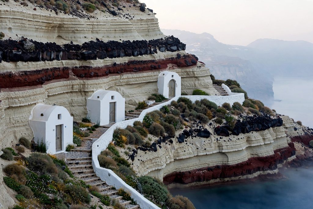 Whitewashed cave houses with vaulted roofs and arched doors carved into the volcanic caldera cliff of Santorini.