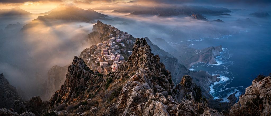 An epic, wide-angle landscape photograph showing the white village of Olympos perched precariously on a jagged mountain spine, high above the distant blue Aegean Sea at twilight.