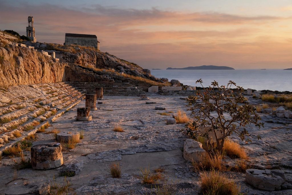 The ruins of the Telesterion hall at Eleusis in low evening light, with rock-cut steps, column bases and the bay beyond.