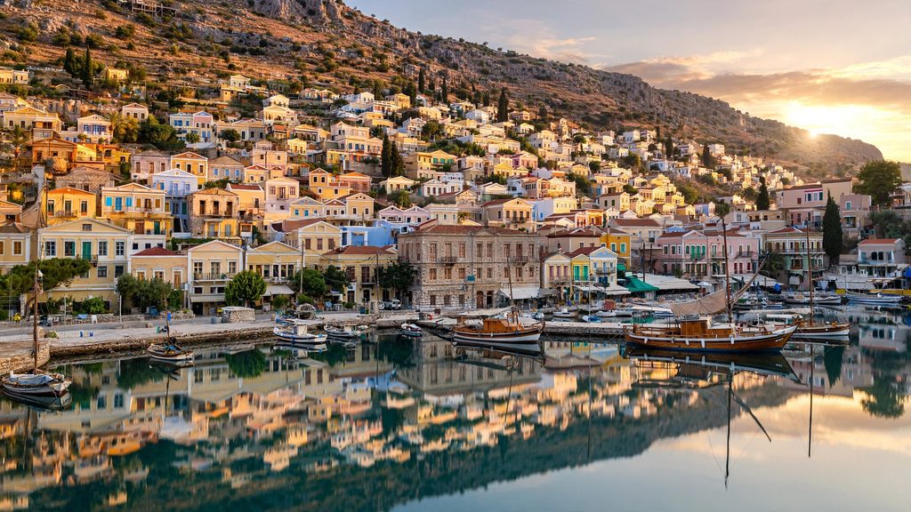 A wide photograph of hundreds of colorful neoclassical Greek mansions climbing a steep harbor hill from turquoise water under golden afternoon sunlight.